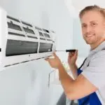Technician in work uniform installing air conditioning unit indoors for AirLux Heating & Cooling HVAC services.