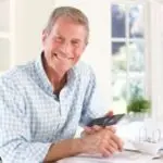 Elderly man in checked shirt holding smartphone, discussing HVAC replacement signs in bright room with notebook.