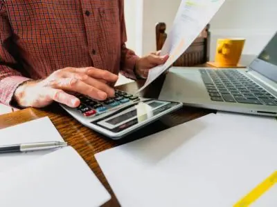 Person calculating energy bills with a calculator, holding documents, and using a laptop, reflecting concerns about rising utility costs related to air conditioning efficiency.