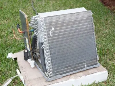 Evaporator coil from an air conditioning system, showing fins and tubing, placed on a cardboard base in a grassy area, illustrating the importance of AC coil cleaning for optimal cooling efficiency.