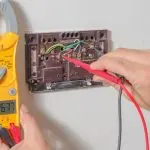 Hands using a multimeter to troubleshoot a thermostat's wiring at Jerry Brittingham A/C & Heat, Inc. for HVAC maintenance.