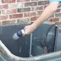 Hand holding a handheld sprayer near a container with wiring against a brick wall in HVAC maintenance setting.