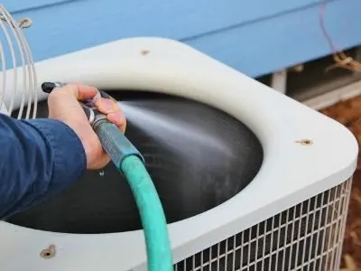 Person cleaning a condenser coil of an air conditioning unit with a hose, emphasizing HVAC maintenance and efficiency.