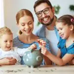 Family happily depositing coins into a large pink piggy bank in a cozy living room, related to financial education.