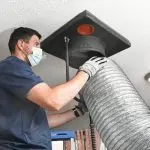 Technician in mask performing HVAC maintenance on a large metallic duct in Daytona Beach home.