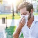 Man in a white shirt outdoors, wiping his nose with a tissue on a sunny day, highlighting HVAC service for allergy sufferers.