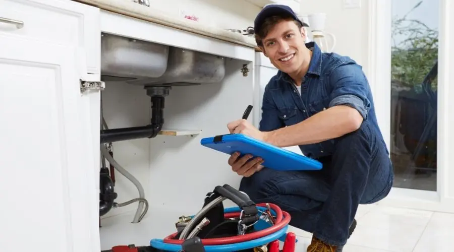 Plumber inspecting under kitchen sink with tools and clipboard, representing plumbing services and maintenance solutions.