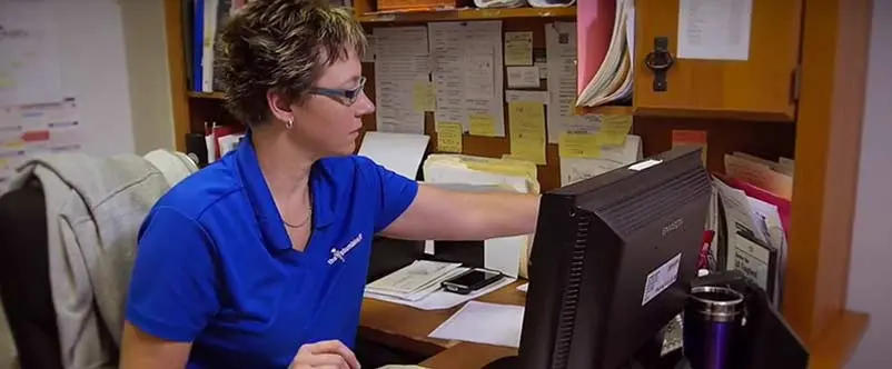 Woman in blue shirt working at desk with computer, surrounded by paperwork and office supplies, representing The Plumbing Dr's customer service and administrative support.