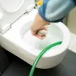 Person using a green hose to flush a toilet, demonstrating plumbing maintenance techniques related to emergency plumbing services.