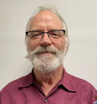 Dr. Charlie, plumbing technician at The Plumbing Dr., smiling with glasses and a beard, wearing a maroon shirt against a neutral background.