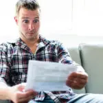 Man sitting on a couch, looking surprised while holding a document, reflecting unexpected plumbing issues or service responses related to The Plumbing Dr's services.