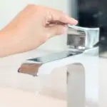 Hand turning on a modern bathroom faucet, illustrating water conservation techniques related to plumbing services.