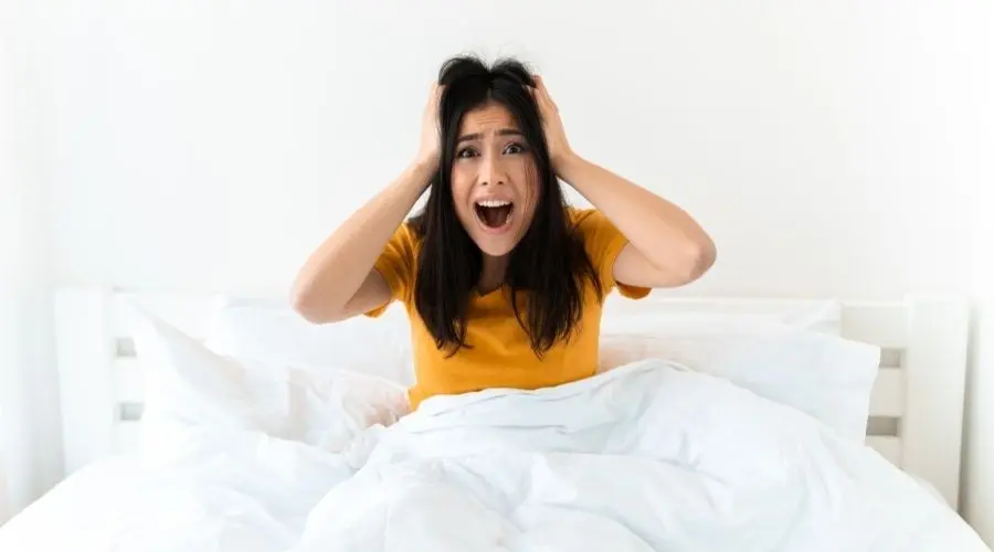 Woman sitting in bed with a distressed expression, holding her head, conveying stress or anxiety, relevant to plumbing emergencies and maintenance tips.