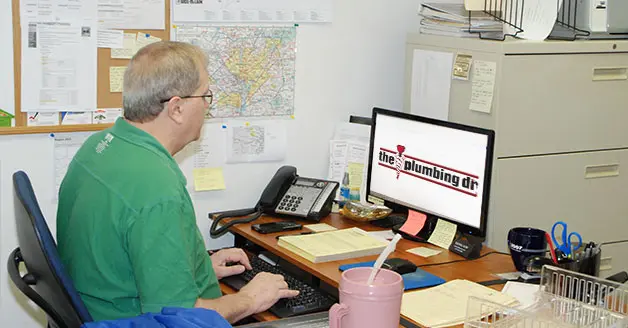 Man working at desk with computer displaying The Plumbing Dr logo, surrounded by office supplies and a map, emphasizing customer service and feedback.