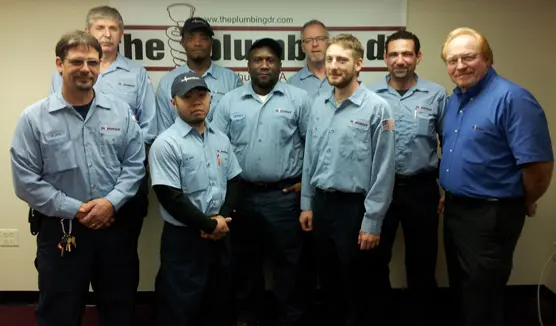Alrington-Plumber-The-Plumber-Doctor-Staff1 Team of plumbers from The Plumbing Dr, wearing blue uniforms, standing in front of a company banner, showcasing their commitment to quality plumbing services in Reston, VA.