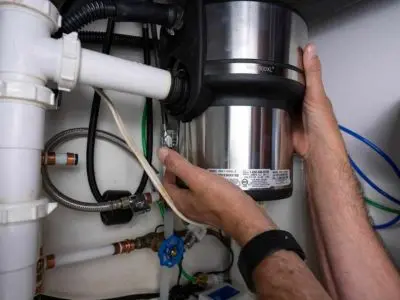 Hands installing a garbage disposal under a kitchen sink, showcasing plumbing connections and maintenance tools, emphasizing professional plumbing services for garbage disposal repair and installation.