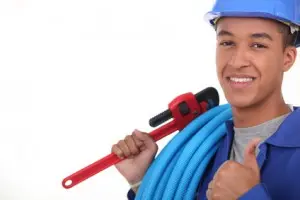 Smiling plumber in a blue uniform and hard hat holding a pipe wrench and plumbing hose, representing plumbing inspection services for new homeowners.