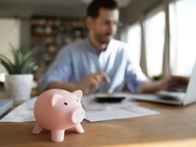 Piggy bank in foreground with a person working on a laptop, symbolizing savings and financial management related to lower utility bills from energy-efficient water heater maintenance.