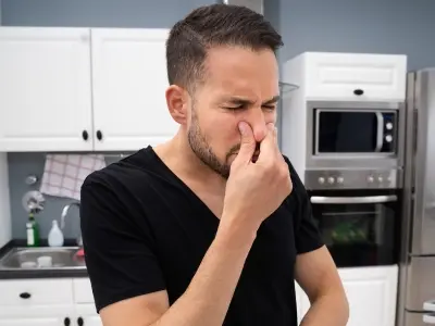 Man holding his nose in a kitchen, indicating foul odors from a garbage disposal, reflecting signs of malfunction and the need for professional plumbing services.