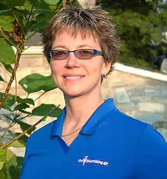 Tracy Bennett, business manager at The Plumbing Dr, wearing a blue polo shirt with the company logo, standing outdoors among greenery, emphasizing her role in scheduling and business efficiency.
