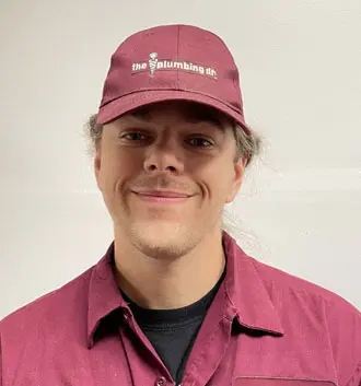 Smiling plumber assistant wearing a maroon cap and uniform from The Plumbing Dr, showcasing a friendly demeanor and professional appearance.