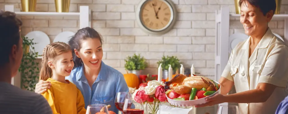 Family gathering around a dining table with a woman serving a platter of food, celebrating Thanksgiving with joy and togetherness.