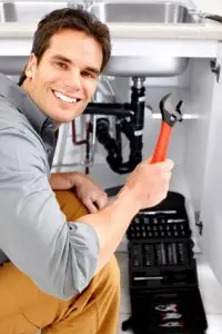 Reasons To Install A Garbage Disposal In Your Home Smiling plumber holding a wrench under a kitchen sink, showcasing plumbing tools and pipes, relevant to garbage disposal installation and repair services.