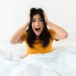 Woman in bed with a shocked expression, hands on her head, conveying surprise or fright, relevant to Halloween bathroom decoration tips.