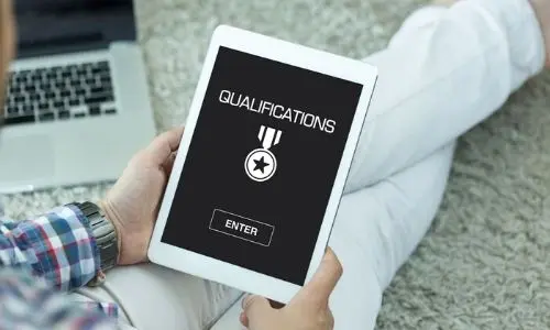 Person sitting on a carpet holding a tablet displaying the word "QUALIFICATIONS" with a medal icon, emphasizing the importance of hiring qualified plumbers.