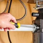 Hand using a screwdriver on a garbage disposal under a sink, highlighting plumbing connections and tools for repairs.