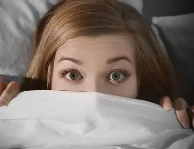 Woman looking scared while hiding under a blanket, representing the fear of spooky sounds in the home, related to furnace issues.