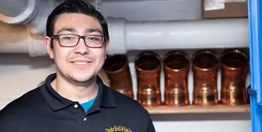 Smiling male employee of Charles Krull & Son Plumbing & Heating in a workshop with copper mugs in background.