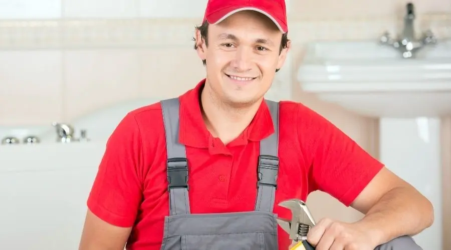 Smiling male plumber in red shirt and gray overalls holding an adjustable wrench in a clean bathroom setting.