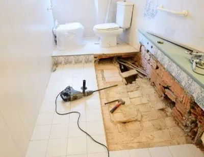 Bathroom remodeling in progress, featuring a partially removed floor exposing plumbing, tools like a hammer and drill, and a toilet in the background, highlighting renovation trends and plumbing upgrades.