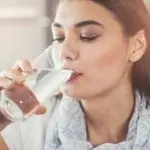 Woman drinking clear water, wearing light scarf; highlights hydration in home water filtration context.