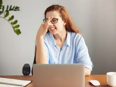 Woman with glasses expressing frustration while sitting at a desk with a laptop, notebook, and coffee cup, illustrating the stress of dealing with plumbing issues like garbage disposal odors.