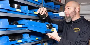 Man in a dark polo shirt organizing labeled blue storage bins in a plumbing and heating service workspace.