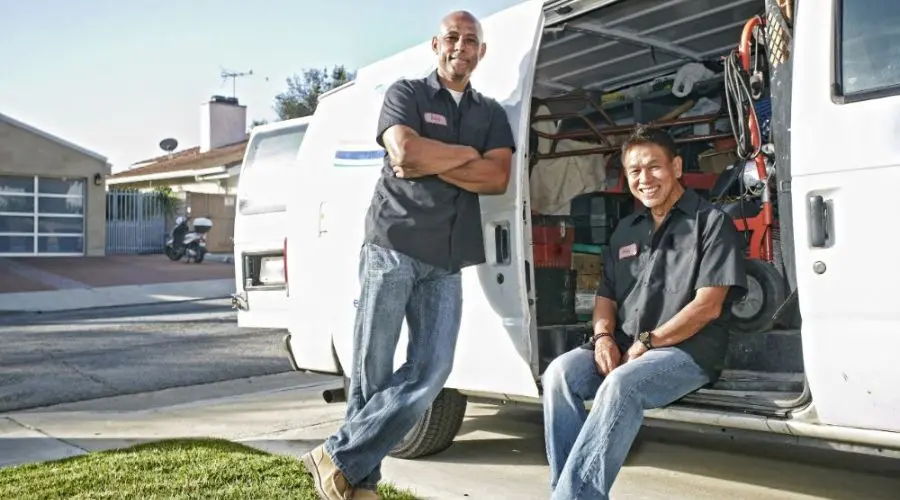 Two plumbers in uniforms smiling next to a service van filled with plumbing tools and equipment, representing Charles Krull & Son, Inc. Plumbing & Heating services.