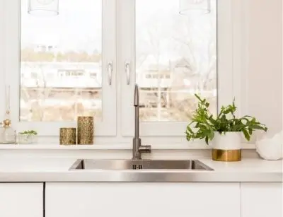 Modern kitchen sink with sleek faucet, natural light from window, decorative plants, and stylish countertop accessories, emphasizing kitchen plumbing upgrades and design trends.