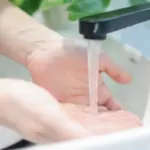 Hands under running water from a faucet in a natural setting, promoting plumbing and heating hygiene tips.