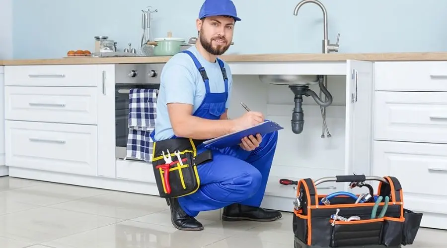 Male plumber in blue overalls writing on a clipboard in a modern kitchen with a toolbox. Charles Krull & Son, Inc.