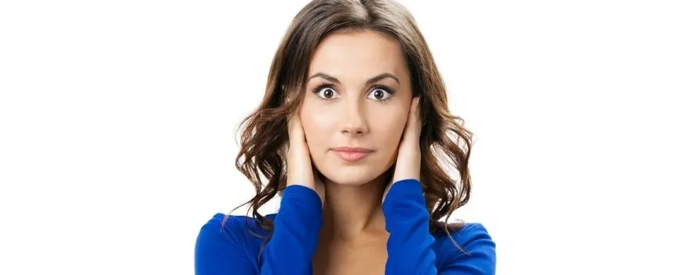 Young woman with dark wavy hair in blue top covering ears, surprised expression, against a plain white background.