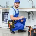 A plumber in blue overalls and cap working at a kitchen sink with tools, representing Charles Krull & Son, Inc.