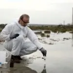 Person in protective gear collecting water samples with tools at an outdoor site for Charles Krull & Son, Inc.