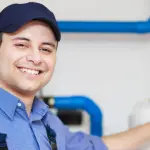 Male plumber in blue cap and shirt smiling, with plumbing tools and blue pipes, focusing on water heater services.