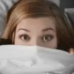 Young woman with wide eyes under a light blanket, showing furnace noise concerns for homeowners at Charles Krull & Son.