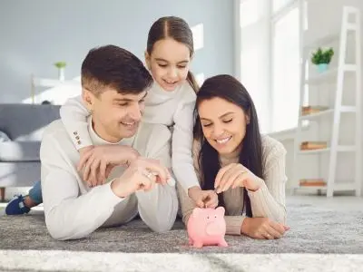 Family saving money together, inserting coins into a pink piggy bank on a living room floor, symbolizing financial planning and energy savings from switching to tankless water heaters.