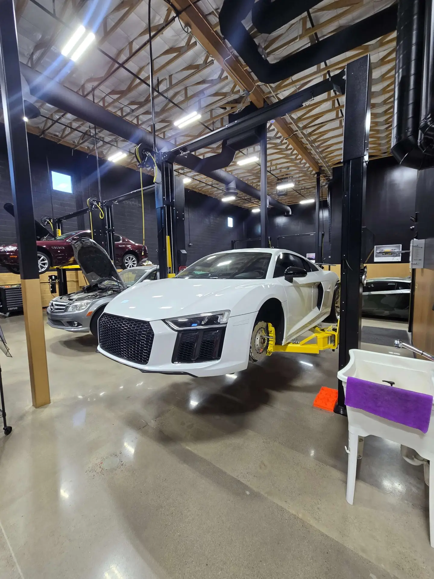 Aces Auto Elite - A white sports car is elevated on a hydraulic lift inside a clean, modern auto workshop. The front wheel is removed, and another car in the background has its hood open. The garage has high ceilings and exposed beams. | Arizona Luxury & Exotic Car Service