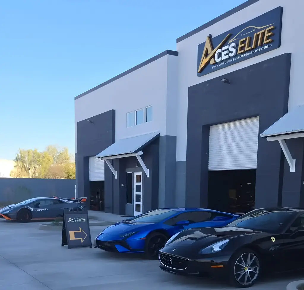 Aces Auto Elite - A modern auto detailing center with a sign reading ACES ELITE and three luxury sports cars—a black Ferrari, a blue Lamborghini, and a black McLaren—parked out front. A signboard with an arrow points to the entrance. | Arizona Luxury & Exotic Car Service