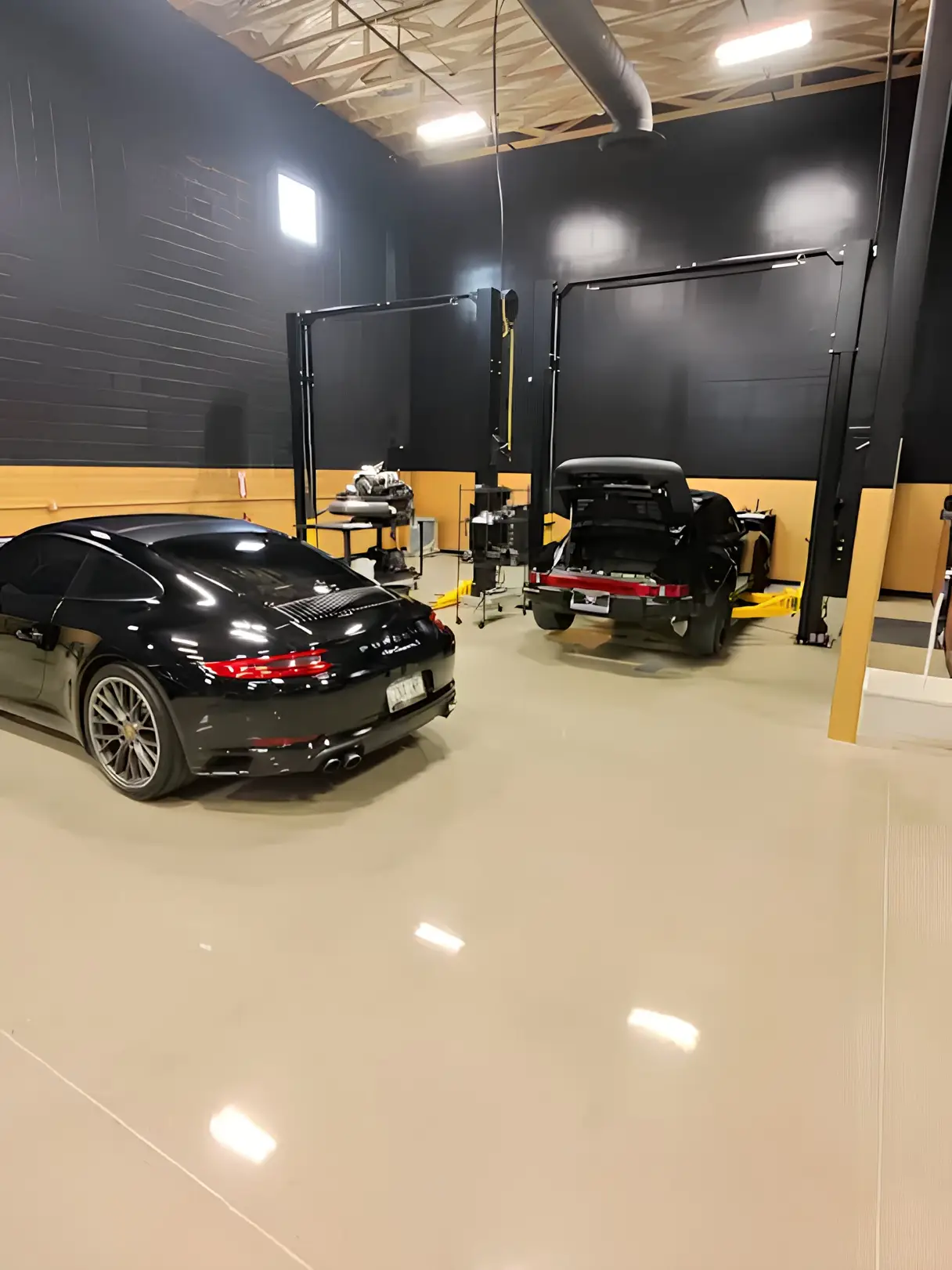 Aces Auto Elite - Two black sports cars in a clean, modern garage; one car is parked with its trunk open on a hydraulic lift, while the other is parked nearby on the shiny beige floor under high ceilings with exposed beams. | Arizona Luxury & Exotic Car Service